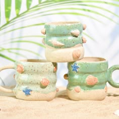 three ceramic mugs with shells and starfish on them are sitting in the sand