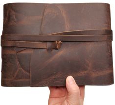 a hand holding a brown leather journal with strap around the edges and clasps on both sides