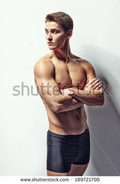 a shirtless young man posing against a white wall with his arms crossed and looking at the camera