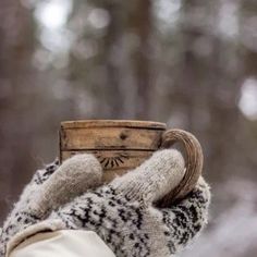 a person wearing mittens holding a cup in their hand with trees in the background