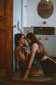 two beautiful young women sitting next to each other on the floor in front of a kitchen