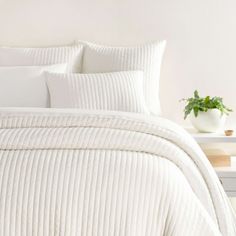 a bed with white comforters and pillows in a room next to a plant on a table