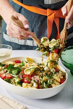 a person holding a wooden spoon over a bowl filled with pasta and veggies