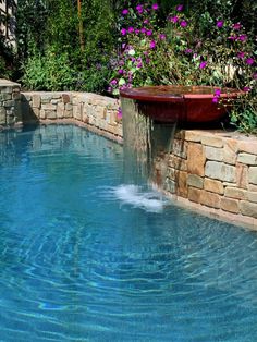 an outdoor pool with water coming out of the side and purple flowers in the background