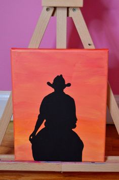 the silhouette of a cowboy on an orange and pink background is mounted to a wooden easel