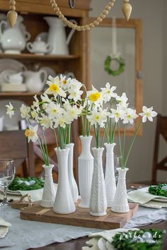 white vases with yellow and white flowers in them on a wooden tray at the dining room table