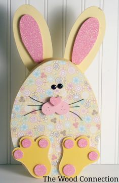 a wooden bunny with pink and yellow feet on it's face, sitting next to a white wall