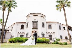 a bride and groom standing in front of a large white building with palm trees on the lawn