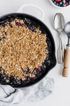 a blueberry crumble in a cast iron skillet next to two bowls of berries and spoons