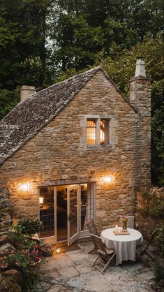 an old stone house with a table and chairs in the front yard, lit up by lights