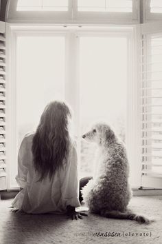 a woman sitting on the floor next to a dog looking out a window with shutters