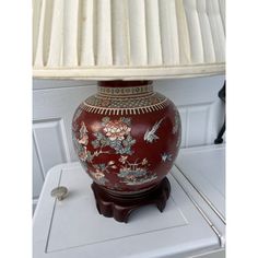 a red vase sitting on top of a white table next to a lamp with a beige shade