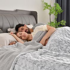 a man and woman laying in bed under a gray comforter with white pillows on it