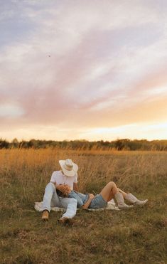 a man and woman sitting on the ground in a grassy field at sunset with clouds overhead