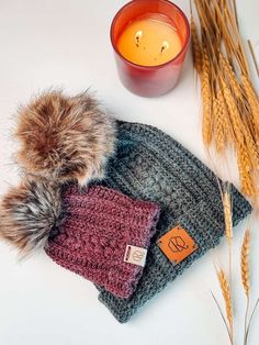 a knit hat with a fur pom sits next to a candle and some wheat stalks