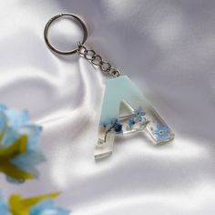 a glass letter keychain with blue flowers in the bottom right corner on a white satin background