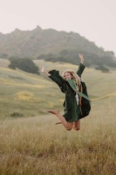 a woman in a green graduation gown jumping into the air with her arms outstretched and legs spread wide