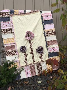 a quilt with flowers on it sitting next to some leaves and plants in front of a building