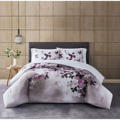 a bed with white sheets and purple flowers on the comforter is in front of a wall