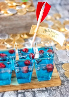 small cups filled with blue liquid and raspberries on a wooden tray next to gold coins