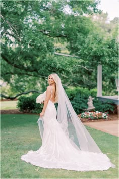 a woman in a wedding dress and veil standing on the grass with her back to the camera