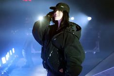 a woman in black jacket and hat standing on stage with lights shining down behind her