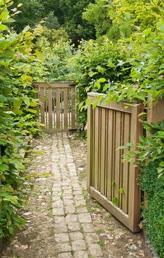 an open wooden gate surrounded by greenery and brick walkway leading to a lush green garden