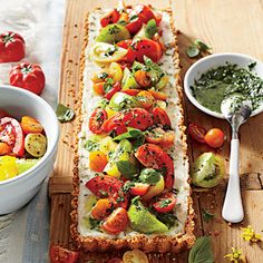 a long rectangular bread with tomatoes, cucumbers and herbs on it next to a bowl of pesto