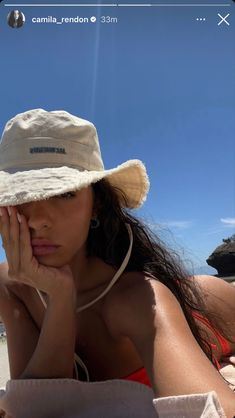 a woman laying on the beach wearing a hat and holding her hand to her face