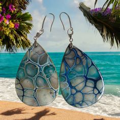 two tear shaped glass earrings sitting on top of a sandy beach next to the ocean