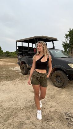 a woman standing in front of a vehicle on a dirt field next to a tree