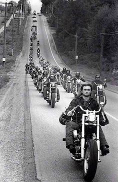 black and white photograph of men riding motorcycles down the road in front of other motorcyclists