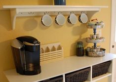 a coffee maker and cups on a shelf in a room with yellow walls, white shelves and wicker baskets