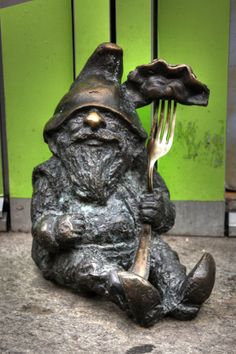 a bronze statue of a gnome holding a fork and sitting on the ground next to a green wall