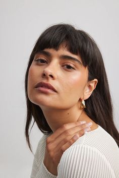 a woman wearing gold earrings and a white sweater looks off to the side with her hand on her shoulder