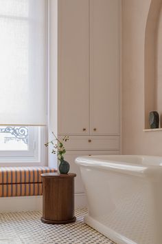 a white bath tub sitting under a window next to a wooden table with flowers in it