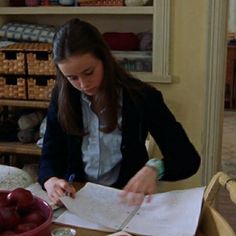 a woman sitting at a table writing on paper with cherries in the bowl next to her