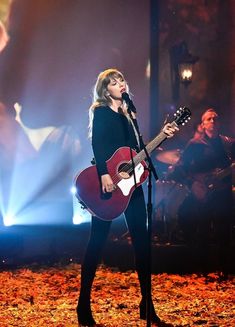 a woman singing into a microphone while holding a guitar in front of her and an audience behind her