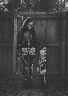 a woman and child dressed up in skeleton costumes, standing next to a wooden fence