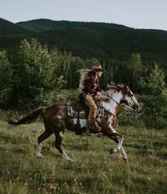 a woman riding on the back of a brown and white horse in a field next to trees