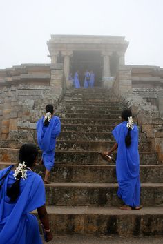 several women in blue dresses are walking up some steps to an ancient structure on a foggy day