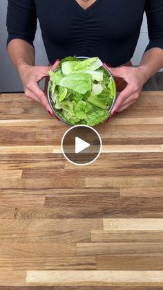 a woman holding a bowl filled with lettuce on top of a wooden table