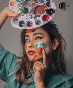 a woman with paint on her face holding a palette over her head and looking at the camera