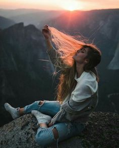 a woman sitting on top of a mountain with her hair blowing in the wind at sunset