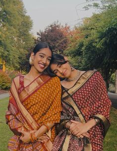 two women standing next to each other in front of some trees and green grass with their arms around one another