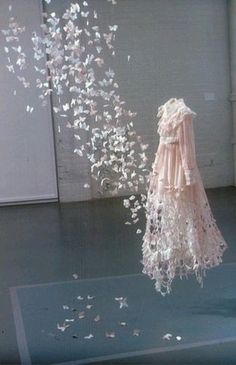 a dress made out of white paper flying in the air next to a wall with flowers on it