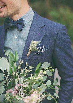 a man wearing a blue suit and bow tie holding a bouquet of greenery in his lapel