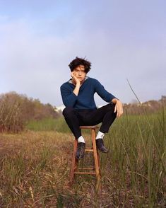 a young man sitting on top of a wooden chair in a field with tall grass