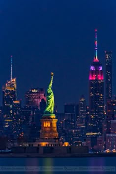 the statue of liberty is lit up at night in front of the city skyline and skyscrapers