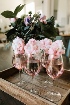 three wine glasses with pink flowers in them on a wooden tray